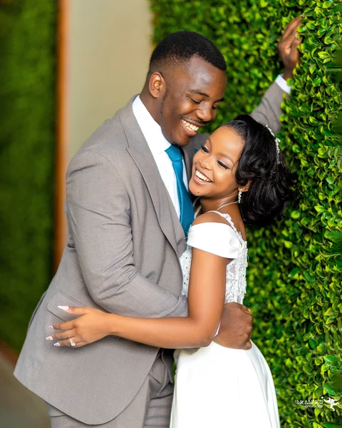 A smiling bride and groom embracing beside a green hedge